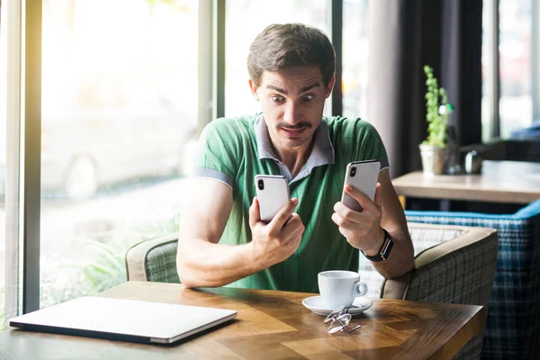 Young shocked busy multiple businessman in green t-shirt looking at smartphone displays in hands with nervous scared face while sitting in cafe, business and freelancing concept