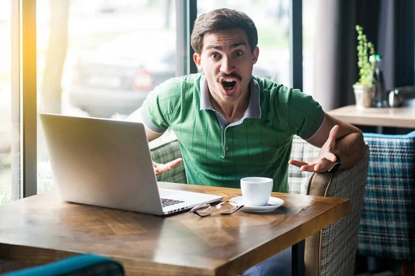 Young angry businessman in green t-shirt  looking and screaming at camera with aggressive crazy face while sitting at table with laptop in cafe, business and freelancing concept