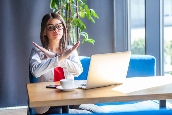 серьезная брюнетка в очках, смотрящая в камеру и показывая поперечный знак, сидя за столом с ноутбуком и кофейной чашкой в кафе, бизнес и фрилансерской концепции
