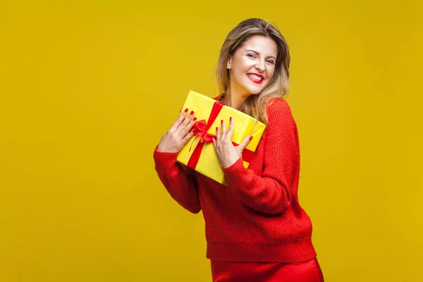 快乐的女人 身穿鲜艳的休闲装 头戴红色口红 拿着包装好的礼品盒 对着镜头笑着 背景是黄色的 生日惊喜 — 图库照片