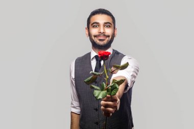 Beyaz gömlekli, yelekli, kırmızı gül çiçekli ve gri arka planda kameraya gülümseyen yakışıklı bir adam.