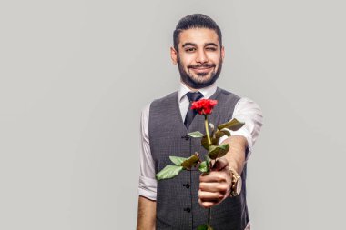 Beyaz gömlekli, yelekli, kırmızı gül çiçekli ve gri arka planda kameraya gülümseyen yakışıklı bir adam.