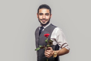 Beyaz gömlekli ve yelekli yakışıklı sakallı adam kırmızı gül çiçeği tutuyor ve gri arka planda gülümseyen kameraya bakıyor.