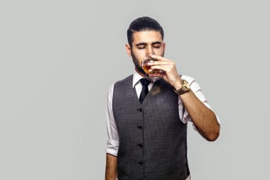 Beyaz gömlekli ve yelekli yakışıklı sakallı adam elinde viski bardağı ve gri arka planda kapalı gözlerle içki içiyor.