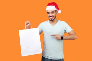 Noel Baba şapkalı ve beyaz tişörtlü neşeli sakallı genç adam ellerinde alışveriş çantasıyla turuncu arka planda dişlek tebessümle kameraya bakıyor.