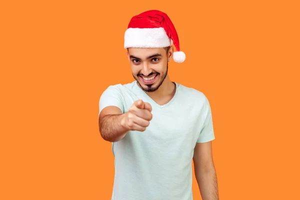 サンタ ハットの若い喜びの髭男とカジュアルな白いTシャツを指差す指とカメラを見てオレンジの背景に笑顔で — ストック写真