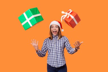 Noel Baba şapkalı, kareli gömlekli, ağzı açık duran esmer kadın turuncu arka planda uçan noel hediyelerini yakalamaya çalışırken ellerini kaldırdı. 