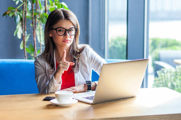 серьезная красивая стильная брюнетка в очках, смотрящая в камеру и показывая предупреждающий жест, сидя за столом с ноутбуком и чашкой кофе в кафе
 