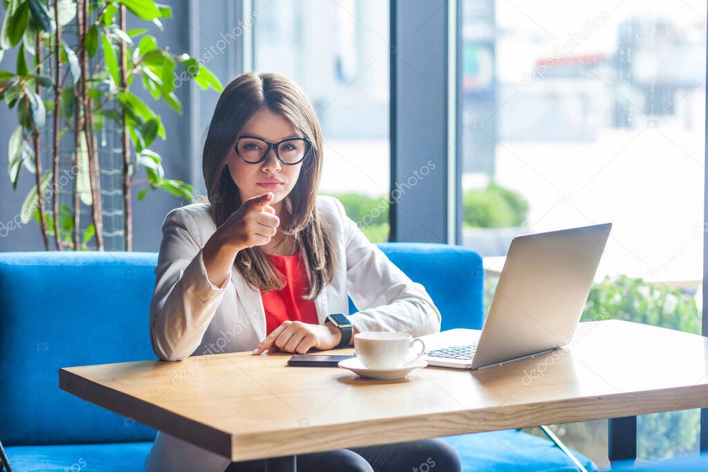 serious beautiful stylish brunette young woman in glasses looking and pointing at camera with serious face while sitting at table with laptop and coffee cup in cafe 