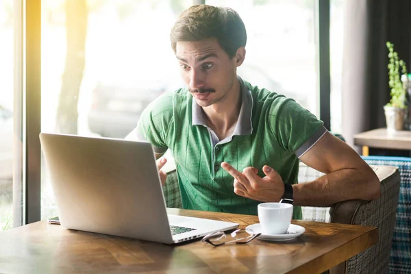 Young angry crazy businessman in green t-shirt looking at laptop screen and showing middle finger fuck signs while sitting at table in cafe