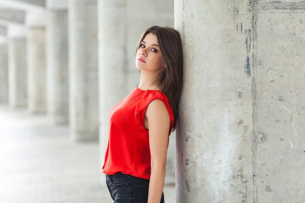 beautiful woman in red blouse and jeans posing while leaning on gray column outdoor