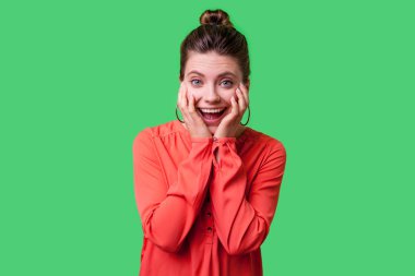 Bun saç stili ve kırmızı bluzlu büyük küpeli hayret bir kadın elleriyle yüzünü tutuyor ve yeşil arka planda şok edici bir ifadeyle kameraya bakıyor.