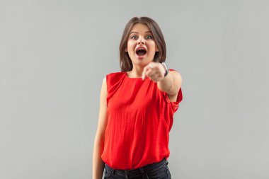 surprised beautiful brunette young woman in red t-shirt pointing and looking at camera with amazed face on gray background