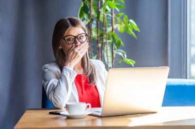 shocked beautiful brunette young woman in glasses looking at camera with big eyes and closing her mouth with hand while sitting at table with laptop in cafe