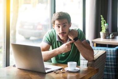 Young serious businessman in green t-shirt looking at camera and pointing at smart watch while sitting at table with laptop in cafe, business and freelancing concept
