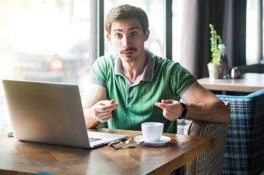 Young businessman in green t-shirt looking at camera and showing money gesture while sitting at table with laptop in cafe, business and freelancing concept