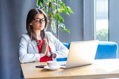 calm beautiful stylish brunette young woman in glasses doing yoga meditation with closed eyes and holding hands in namaste pose while sitting at table with laptop in cafe