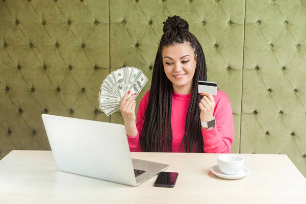 Cash or credit card? Happy beautiful young businesswoman with black dreadlocks hairstyle in pink blouse sitting, holding many dollars, choosing plastic card or cash money, looking at laptop,indoor