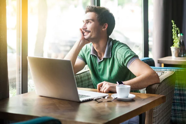 Young smiley happy businessman in green t-shirt looking outside while sitting at table with laptop in cafe, business and freelancing concept