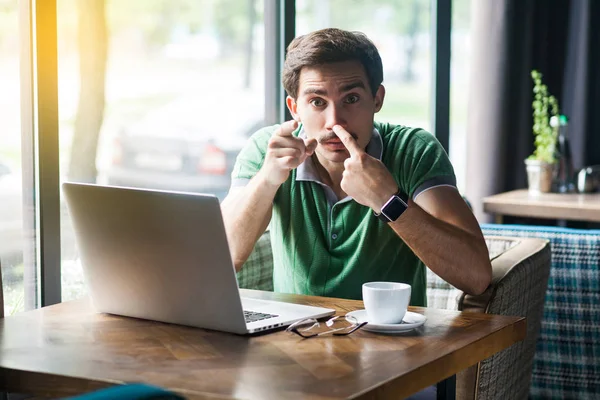Young angry businessman in green t-shirt pointing at camera and touching his nose while showing lie gesture and sitting at table with laptop in cafe, business and freelancing concept