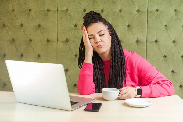 Overwork. Portrait of tired kind beautiful young freelancer woman with black dreadlocks hairstyle in pink blouse is sitting in cafe and fall asleep from fatigue on hand. Indoor, healthy lifestyle