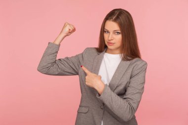 İş elbisesi içindeki kendine güvenen hırslı genç bir kadının portresi gururla pazıları işaret ederek el kaldırıyor, gücü hissediyor, kadın gücü, haklar ve feminizm kavramı. Kapalı stüdyo çekimi, izole.