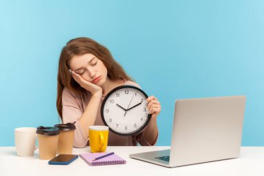 Geç saatlere kadar çalışan yorgun bir ofis çalışanı. Kahve fincanlarıyla çevrili yorgun bir kadın uyuyor ve saati tutuyor, yorgunluk hissediyor, fazla mesai stresi yaşıyor. Mavi arka planda kapalı stüdyo çekimleri