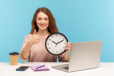 İyi iş, çalışma zamanı! Olumlu, mutlu bir kadın, işyerinde oturan tatmin olmuş bir ofis çalışanı, büyük bir saat tutuyor ve işaret ediyor, tıpkı bir jest gibi. Mavi arka planda kapalı stüdyo çekimleri