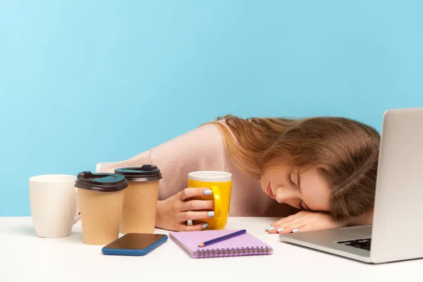 Exhausted fatigued woman office employee sleeping at workplace, lying surrounded by coffee cups, dreaming, resting after tiresome shift, overtime work. indoor studio shot isolated on blue background