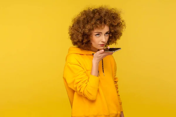 Virtual voice assistant on smartphone. Portrait of curly-haired woman in urban style hoodie giving command to mobile device in her hand, recording message. studio shot isolated on yellow background