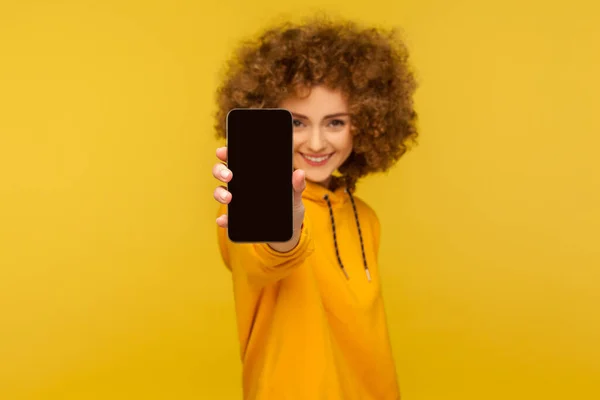 空のディスプレイで携帯電話を示す魅力的な笑顔で陽気な巻き毛の女性の肖像画 アプリ広告のためにモックアップ モバイルWebサービス 黄色の背景に隔離された屋内スタジオで — ストック写真