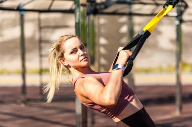 Kapalı alanda antrenman yapan güçlü sarışın bir sporcu, trx süspansiyon askılarını çekiyor, kas yapısı için üst vücut egzersizleri yapıyor, yağ yakıyor, kardiyo gelişimi gösteriyor. Sağlık ve spor faaliyeti kavramı