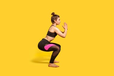 Spor kıyafetleri içinde, sıkı spor kıyafetleri giymiş atletik bir kadın, vücut sporu egzersizi yapıyor, dengesini koruyor, ısınıyor ve kaslarını çalıştırıyor. Tam uzunluktaki stüdyo çekimi, sarı arka planda izole edilmiş.