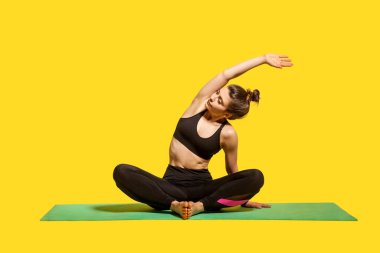 Sıska bir kadın, sıkı spor kıyafetleri içinde nilüfer pozisyonunda oturmuş yoga yapıyor, kaldırılmış ellerle yan yana eğiliyor, esneme egzersizleri yapıyor. Stüdyo çekimi sarı arkaplanda izole edildi