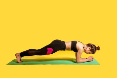 Düşük kalas pozisyonu. Sıska kız, dar spor kıyafetleriyle yoga yapıyor, Ardha Phalakasana egzersizi yapıyor, kaslarını çalıştırıyor. Stüdyo çekimi, spor çalışmaları sarı arka planda izole edilmiş.