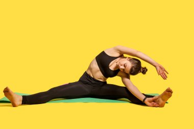 Spor jimnastikçi kadın, dar spor kıyafetleriyle mindere oturmuş, bacakları açık, ayak parmaklarına dokunmak için eğiliyor, geriye ve el kaslarına yaslanıyor, esneklik egzersizleri yapıyor. Stüdyo çekimi, sarı renkte.