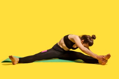 Sıska jimnastikçi kadın dar spor kıyafetleriyle mindere oturmuş, bacakları açık, ayak parmağına dokunmak için eğiliyor, kas egzersizleri yapıyor, esneklik egzersizleri yapıyor. Stüdyo çekimi, sarı renkte.