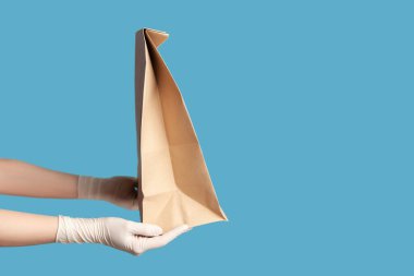 Profil yan görüntüsü, insan elinin beyaz cerrahi eldivenlerle yakın plan görüntüsü. Ellerinde yiyecek ve kağıt paketi var. Güvenli yiyecek teslimatı. İçerde, stüdyoda, mavi arka planda izole edilmiş..