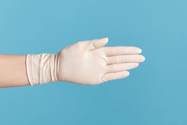 Profil yan görüntüsü, insan elinin beyaz cerrahi eldivenlerle birbirine dokunuşunu ve selamlaşmasını gösteriyor. İçerde, stüdyoda, mavi arka planda izole edilmiş..