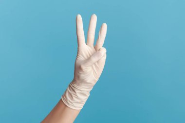 Profil yan görüntüsü, insan elinin beyaz cerrahi eldivenli hali, üç numaranın elleri olduğunu gösteriyor. İçerde, stüdyoda, mavi arka planda izole edilmiş..