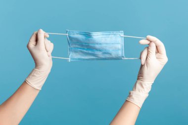 Mavi cerrahi maske takan beyaz eldivenli insan elinin profil yan görüntüsü. İçerde, stüdyoda, mavi arka planda izole edilmiş..