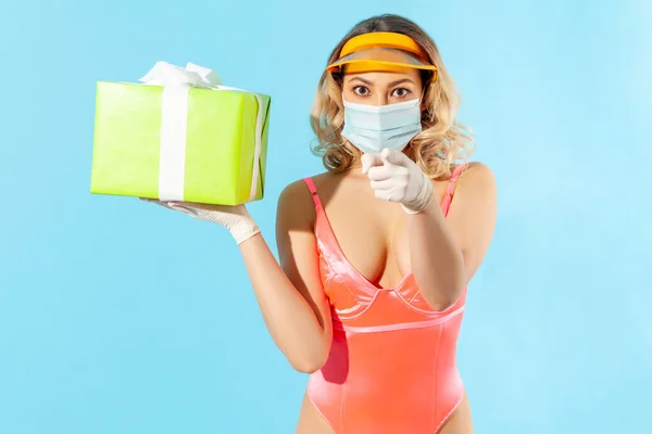 Hey, this present for you! Woman in swimsuit, hygienic face mask and protective gloves, holding gift box and pointing finger to camera, safe birthday celebration in coronavirus covid-19 quarantine