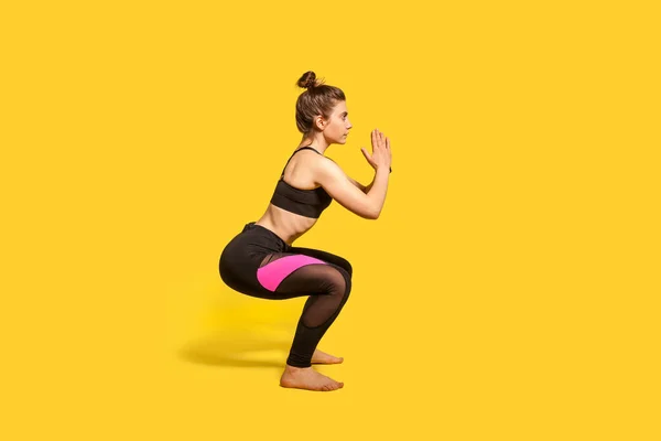 身披发髻的女运动员穿着紧身运动服 蹲着做运动 做下半身运动 保持平衡 锻炼肌肉 全长演播室拍摄 孤立在黄色背景 — 图库照片