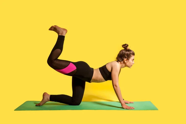 Slim fitness woman with hair bun in tight sportswear practicing yoga, doing one legged plank pose bow leg, training muscle flexibility. studio shot, sport daily workouts isolated on yellow background