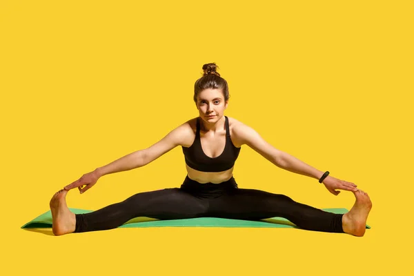 タイトなスポーツウェアで髪パンと体操の女性は 広がった足でマットの上に座って 足の筋肉を伸ばすつま先に触れ スポーツの柔軟性の演習を行う 黄色の背景に隔離された屋内スタジオで — ストック写真