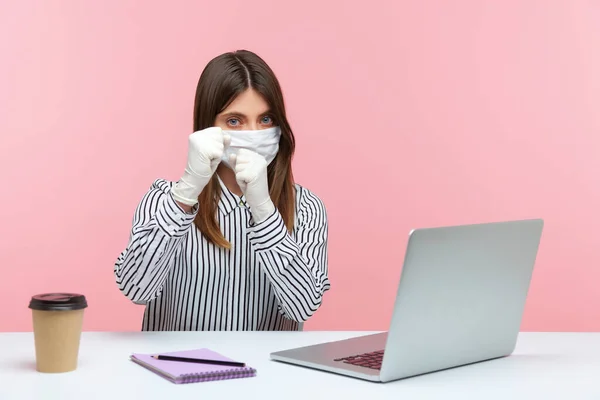 Fight coronavirus. Woman office employee sitting safe healthy with hygienic face mask and protective gloves during quarantine and keeping fists clenched to punch, boxing gesture. indoor, studio shot