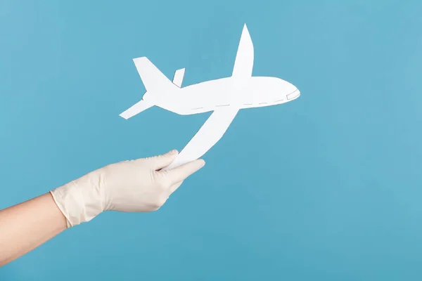 Profile side view closeup of human hand in white surgical gloves holding airplane paper. indoor, studio shot, isolated on blue background.