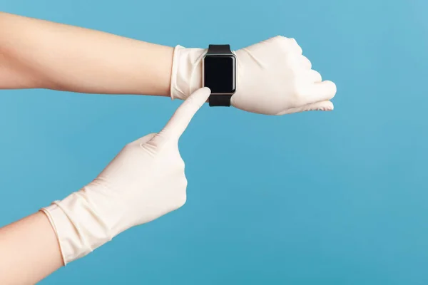 Profile side view closeup of human hand in white surgical gloves holding and showing wirst smart watch and pointing at empty screen. indoor, studio shot, isolated on blue background.