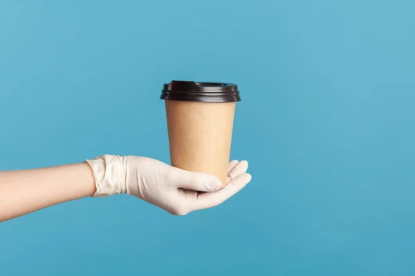 Profile side view closeup of human hand in white surgical gloves holding and showing cup of hot takeaway mug drink in hand. indoor, studio shot, isolated on blue background.