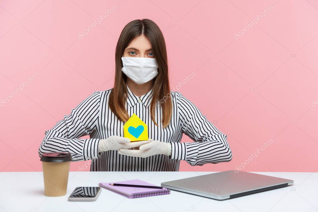 Renting during coronavirus quarantine. Woman real estate agency sitting at workplace, wearing hygienic mask and protective gloves, holding paper house, offering help in mortgage issues, home insurance